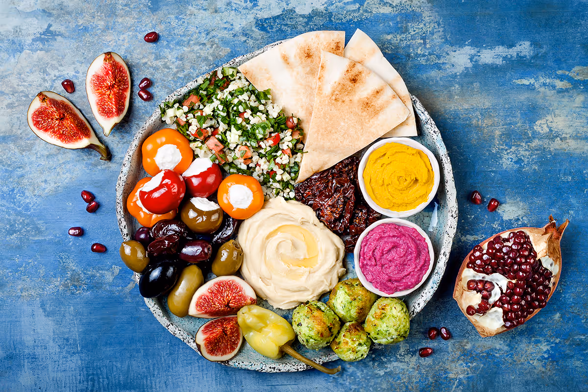 Vorspeisen-Teller mit Oliven, gefüllten kleinen Paprika, Hummus, Tabouleh, Fladenbrot, Falafel, zwei Dips und frischen Feigen auf blauem Hintergrund.