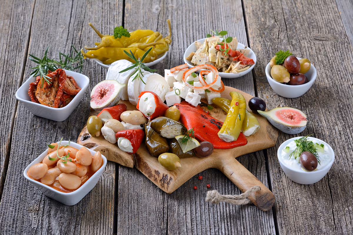 Holzbrett mit gefüllten Paprikaschoten, Oliven, Käse, Feigen, verschiedenen eingelegten Gemüsesorten und kleinen Schalen mit Bohnen, getrockneten Tomaten und Dip auf rustikaler Holztischplatte.